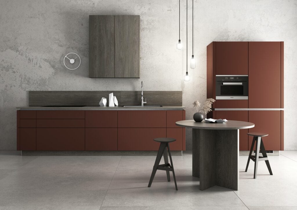 A modern kitchen with minimalist design features red-brown cabinets with lacquered fronts from Bauformat, a wall-mounted clock, a built-in oven, and a small round dining table with two stools. Hanging lights add a contemporary touch.