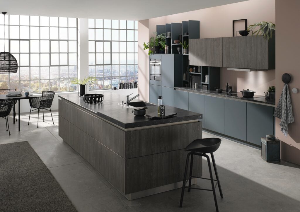 Modern kitchen with grey melamine cabinets, a large island with a black countertop, and a dining area in the background. Large windows allow natural light to fill the space.